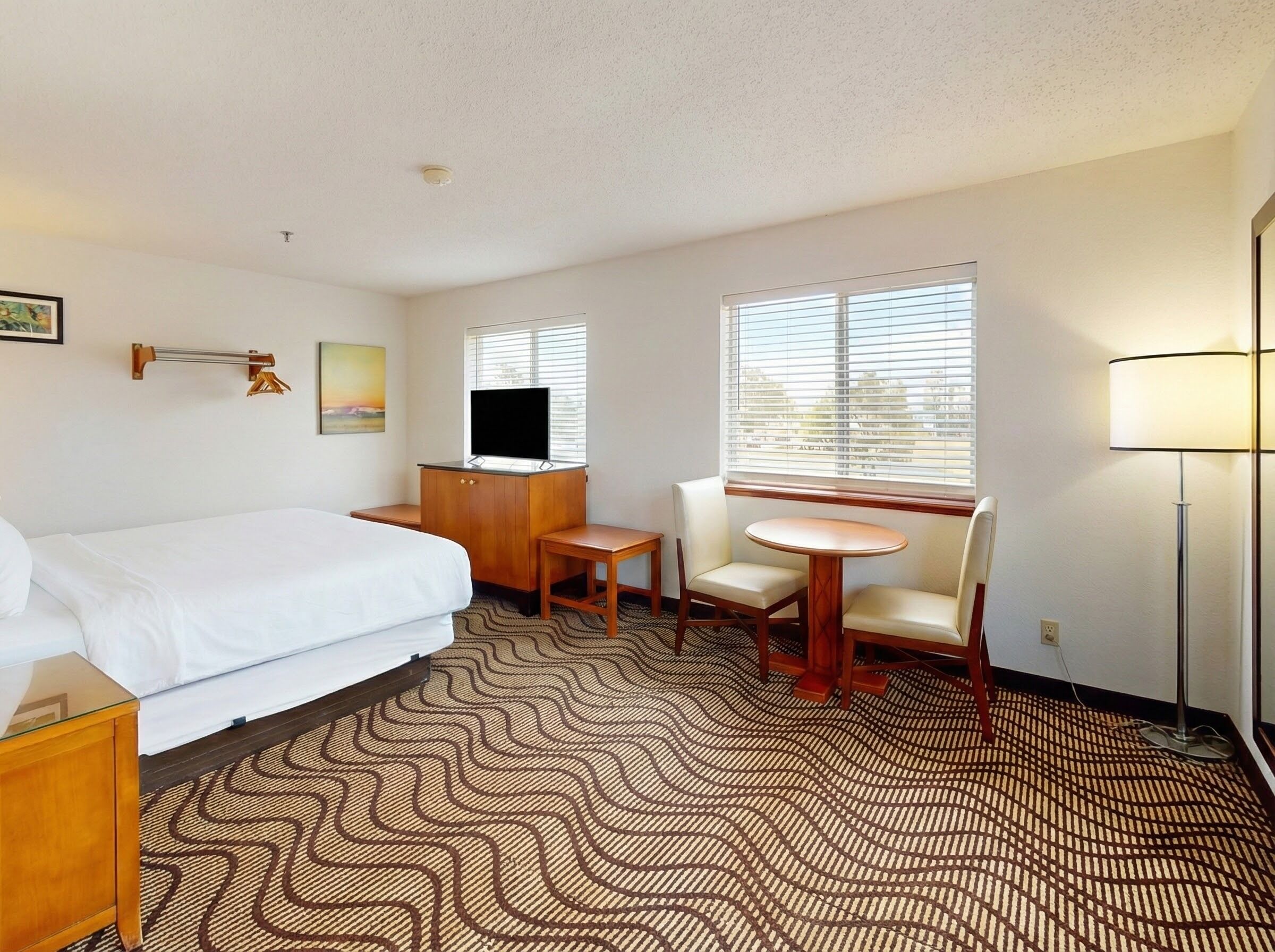 A hotel room with a bed, a desk with a TV, a chair, a small table, and a lamp.
