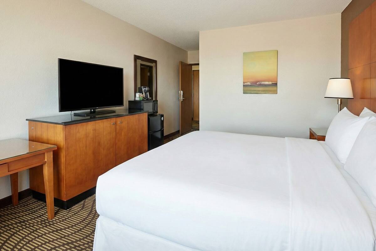 A hotel room with a large bed, a flat-screen TV, a small kitchenette, and a painting on the wall.