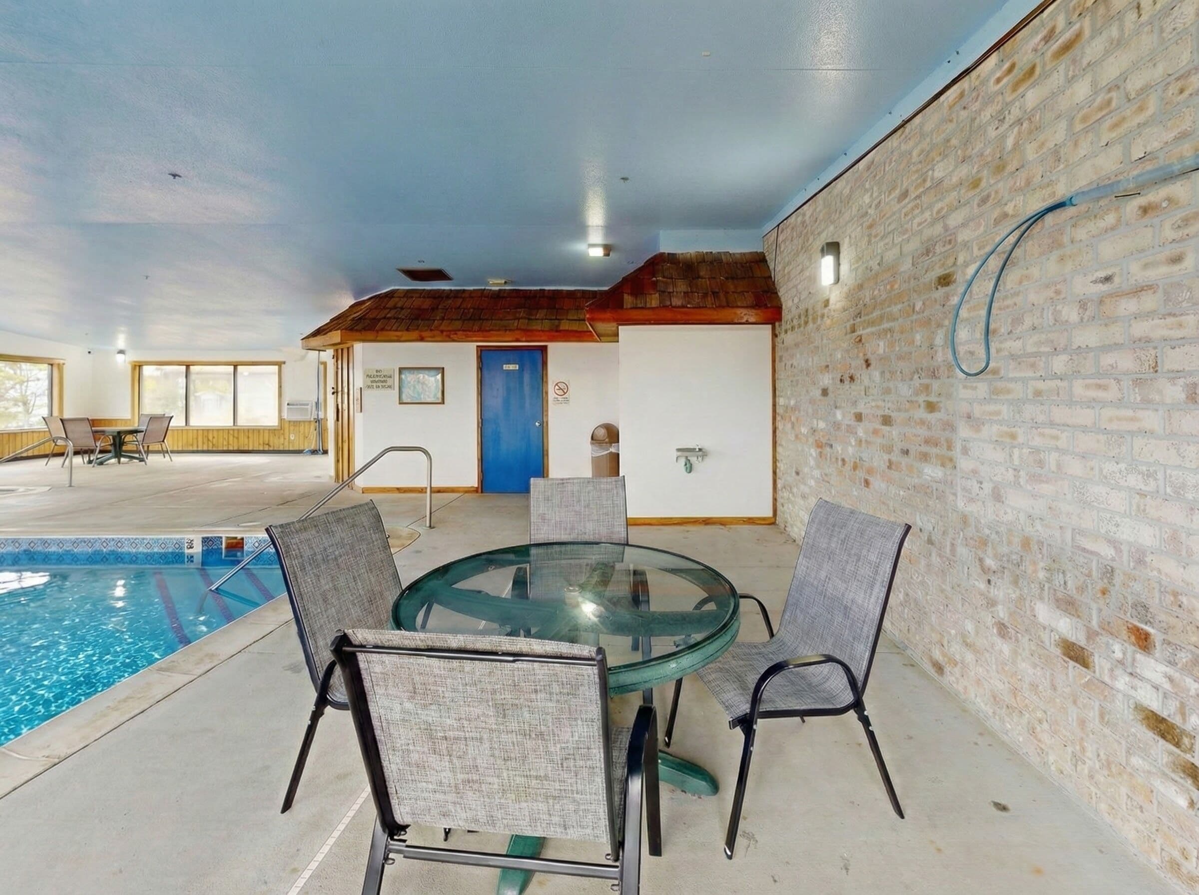Un área de piscina interior con una mesa y sillas de vidrio, una puerta azul y una pared de ladrillo.