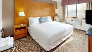 Habitación de hotel con una cama grande, una mesita de noche con lámpara, una silla, un televisor y una ventana con cortinas.