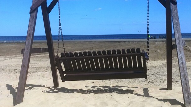 Un columpio de madera en una playa arenosa, con un cielo azul despejado y el océano al fondo.
