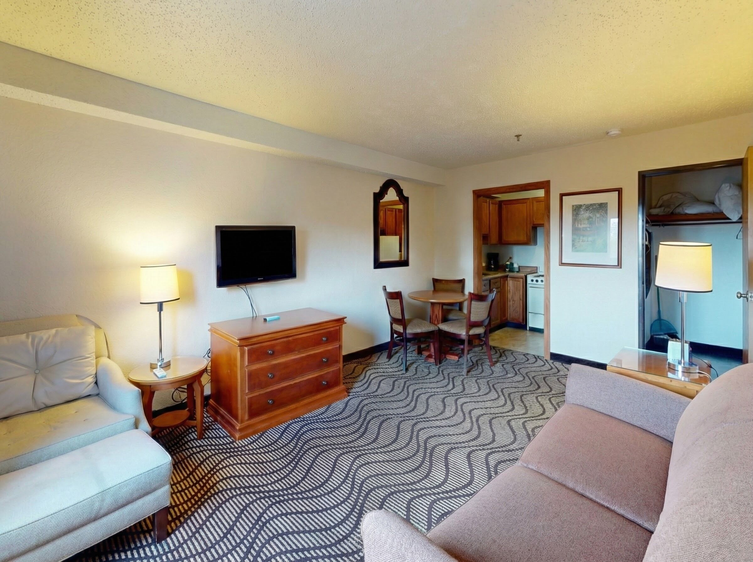 A hotel room with a sofa, a TV, a dresser, a dining table with chairs, a kitchen area, and a patterned carpet.