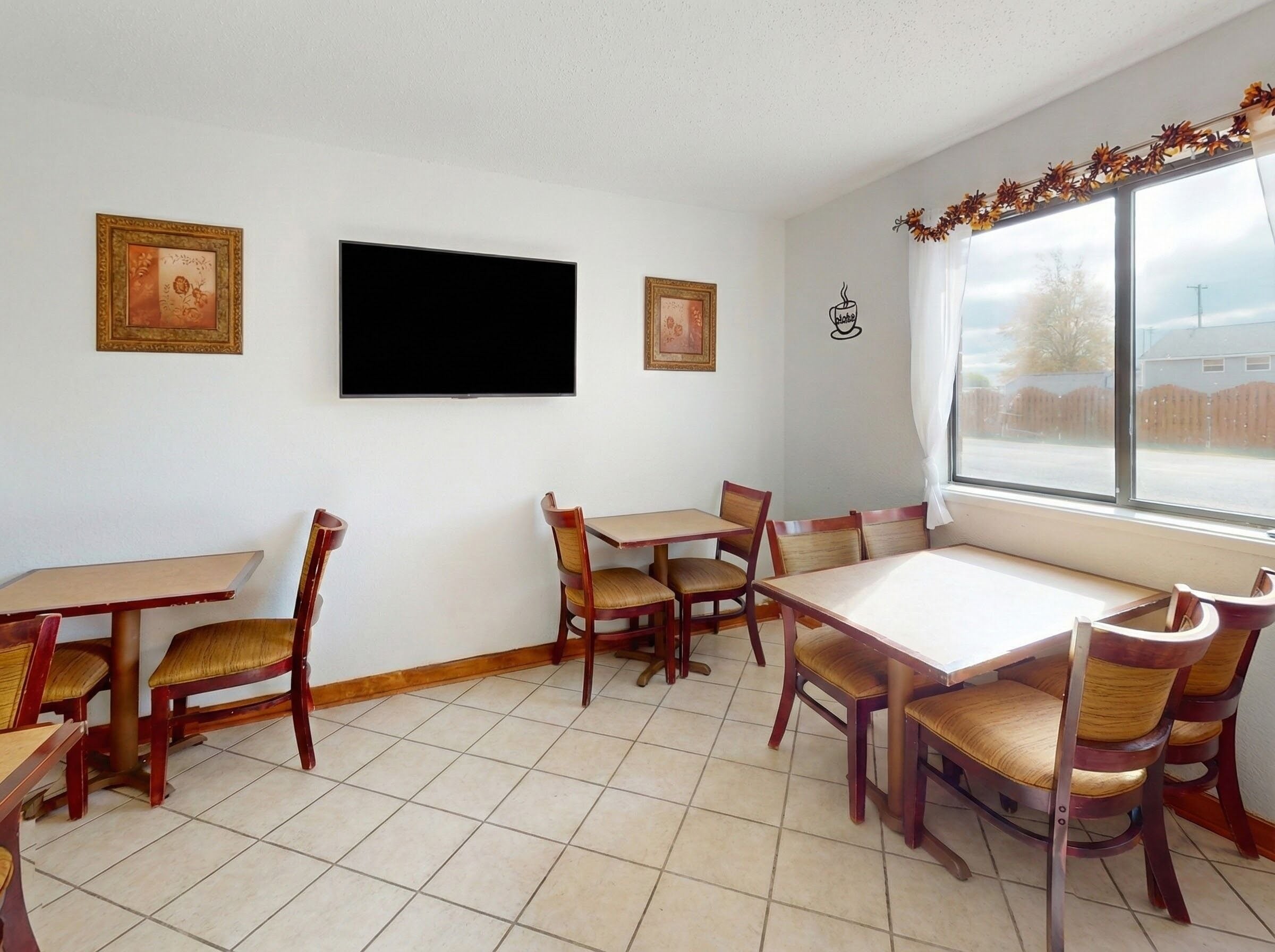 A dining area with wooden tables and chairs, a flat-screen TV, framed pictures on the wall, and a window with a view of the outside.