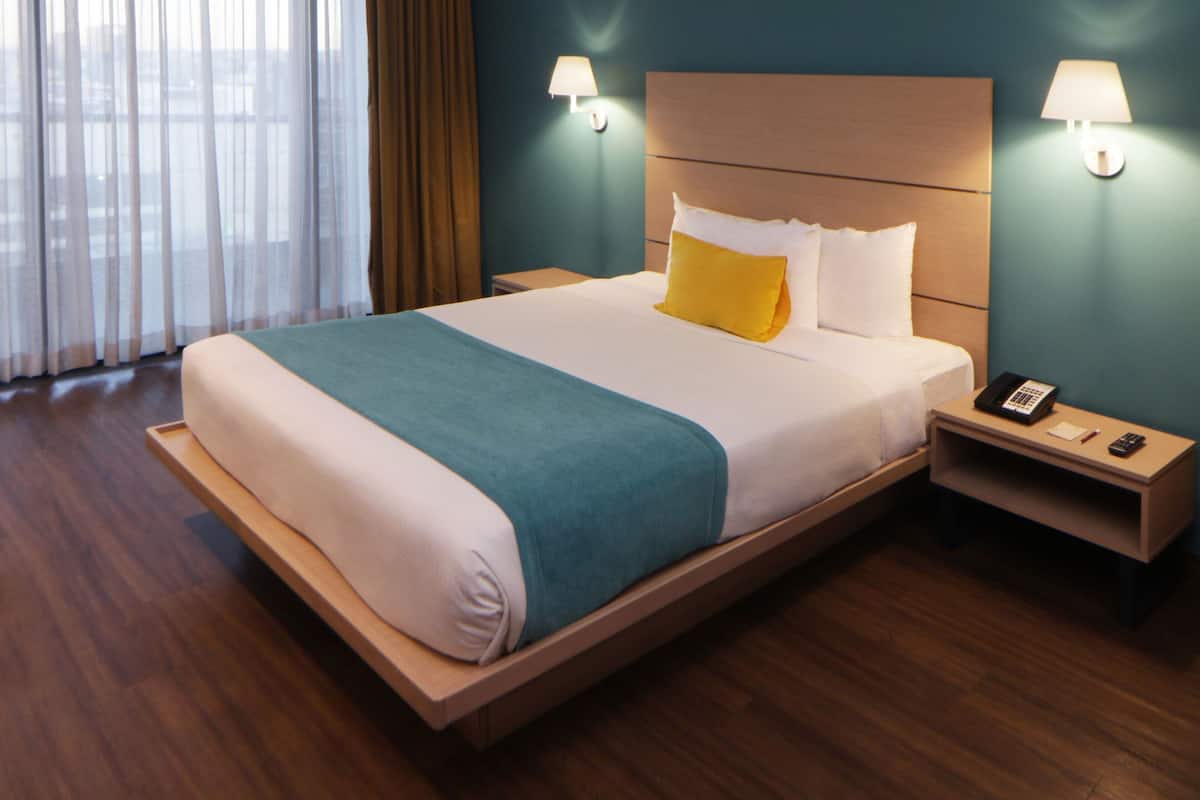 A hotel room with a large bed, two bedside lamps, a nightstand with a phone, and a view through sheer curtains.