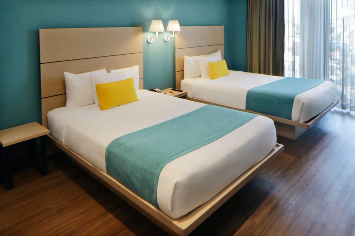 A hotel room with two beds, each with white linens and teal accents, wooden headboards, and bedside tables.