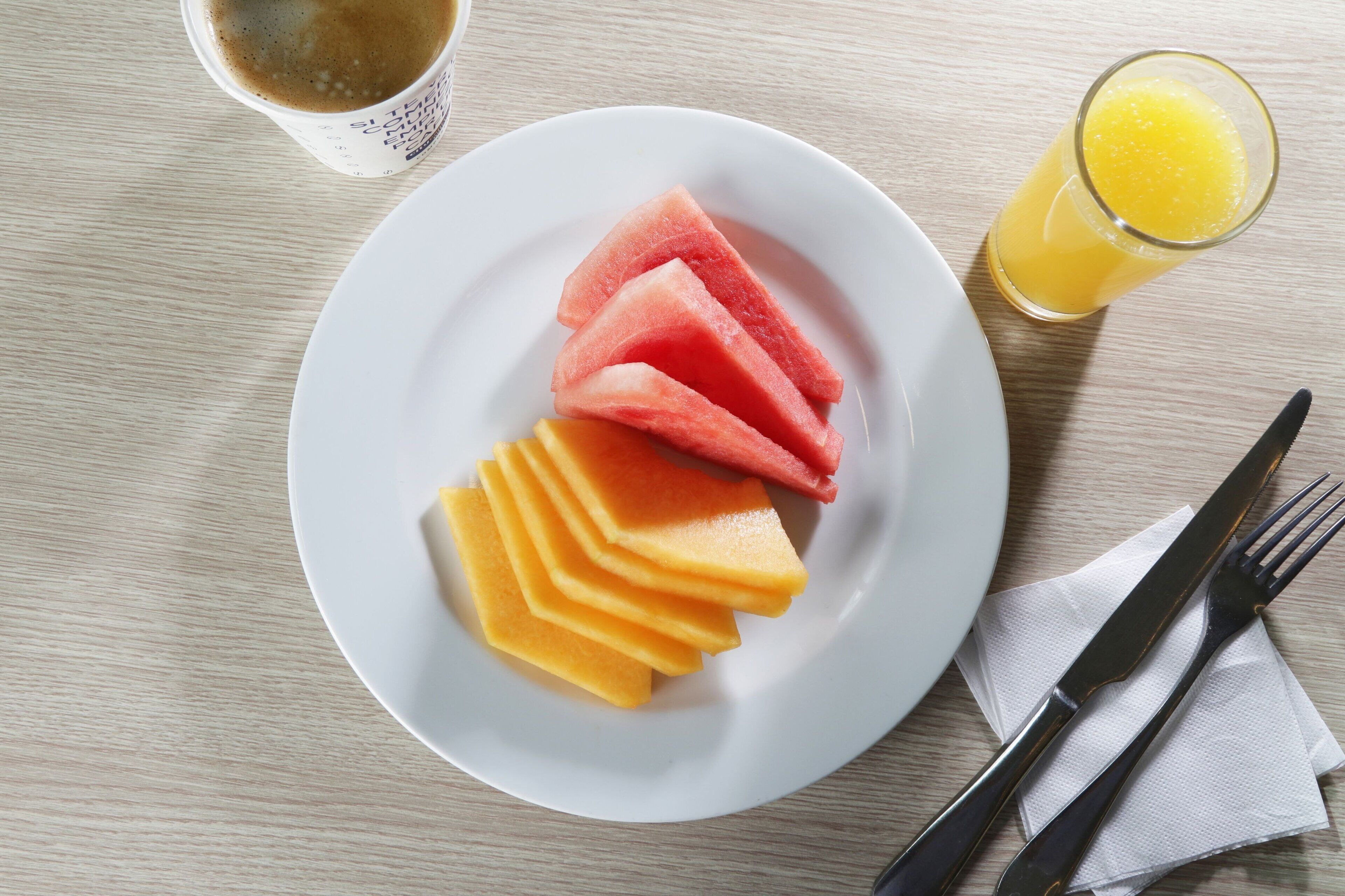 A plate with watermelon and cantaloupe slices, a glass of orange juice, and a cup of coffee on a wooden table.