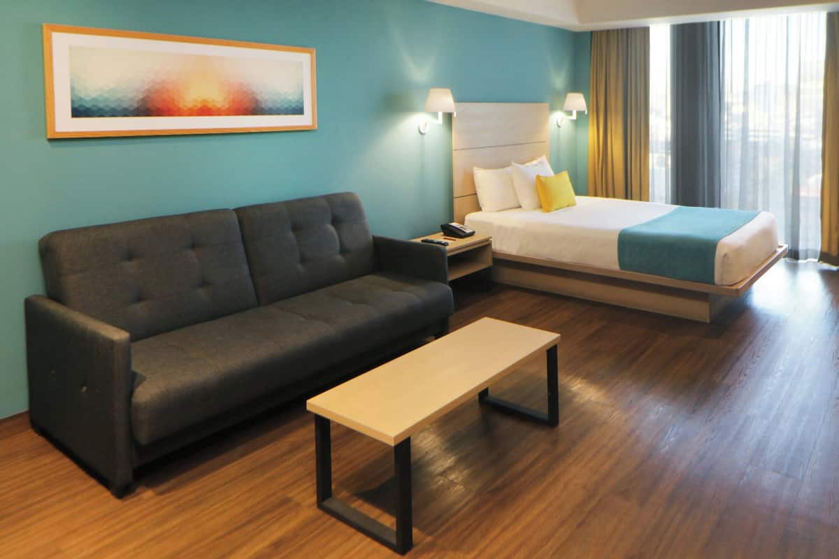 A hotel room with a sofa, a bed with a blue blanket, a bedside table, and a painting on the wall.