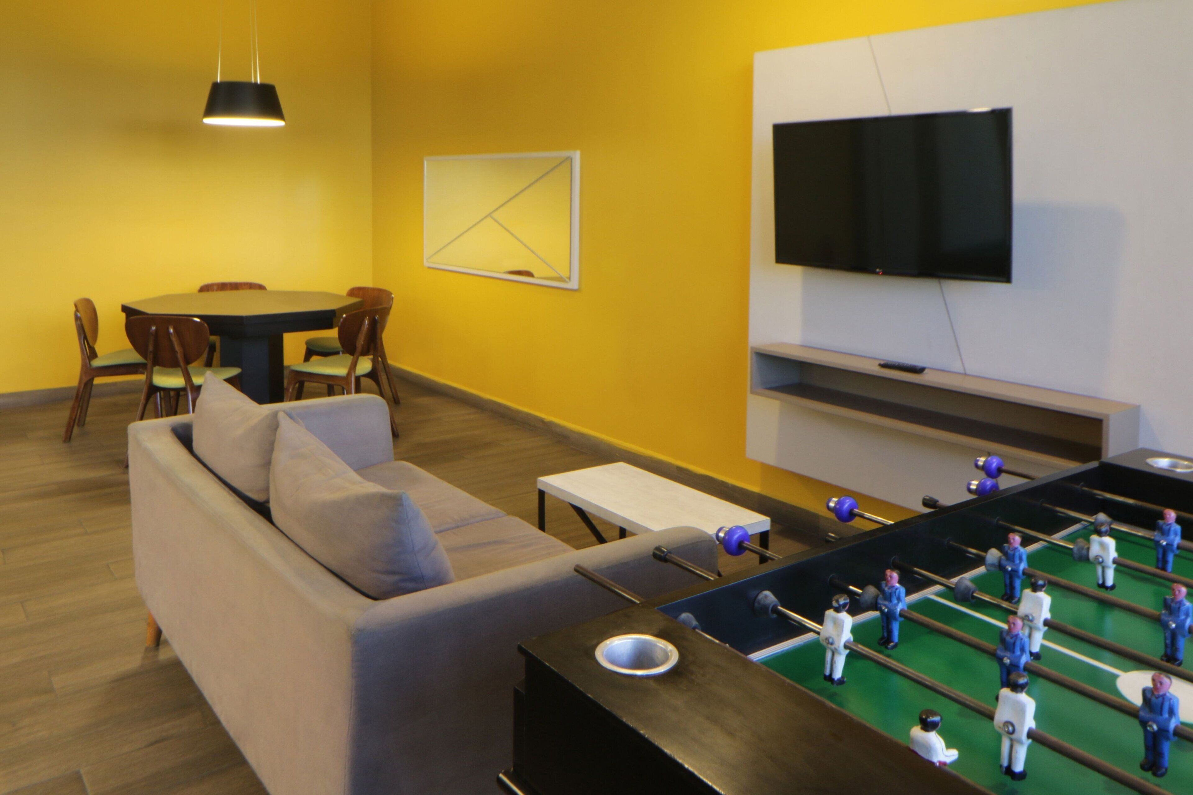 Uma sala de estar com mesa de pebolim, sofá, mesa de jantar com cadeiras e uma TV de tela plana fixada na parede.