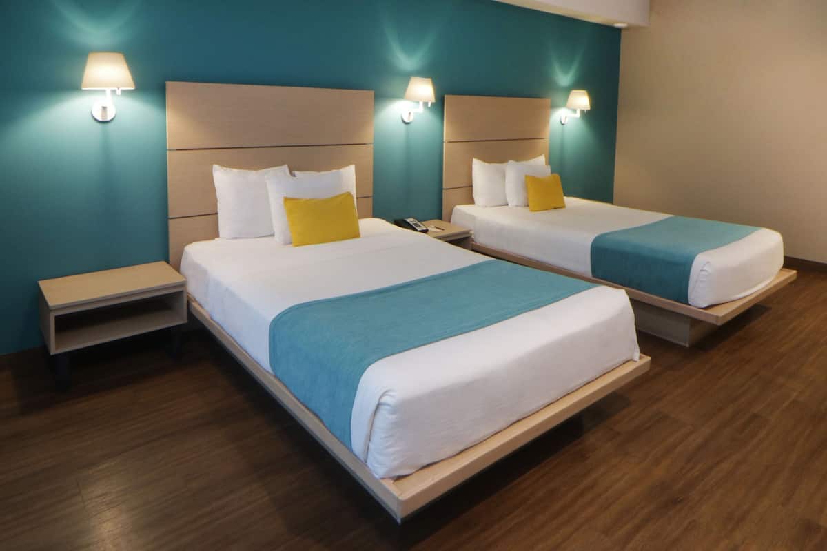 A hotel room with two beds, each with a blue and white bedspread, a wooden headboard, and a nightstand with a lamp.