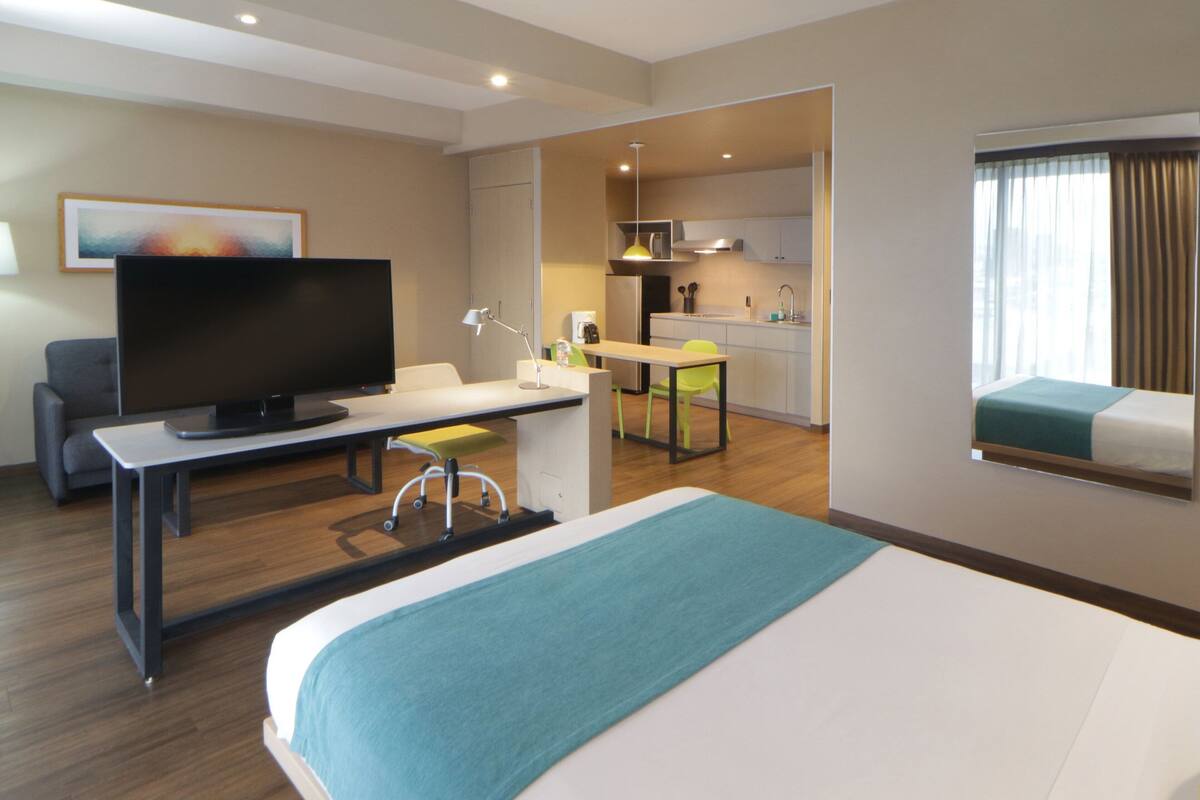 A hotel room with a bed, a desk with a television, a chair, a small kitchen area, and a window with curtains.