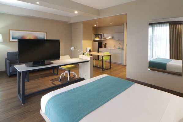 A hotel room with a bed, a desk with a television, a chair, a small kitchen area, and a window with curtains.