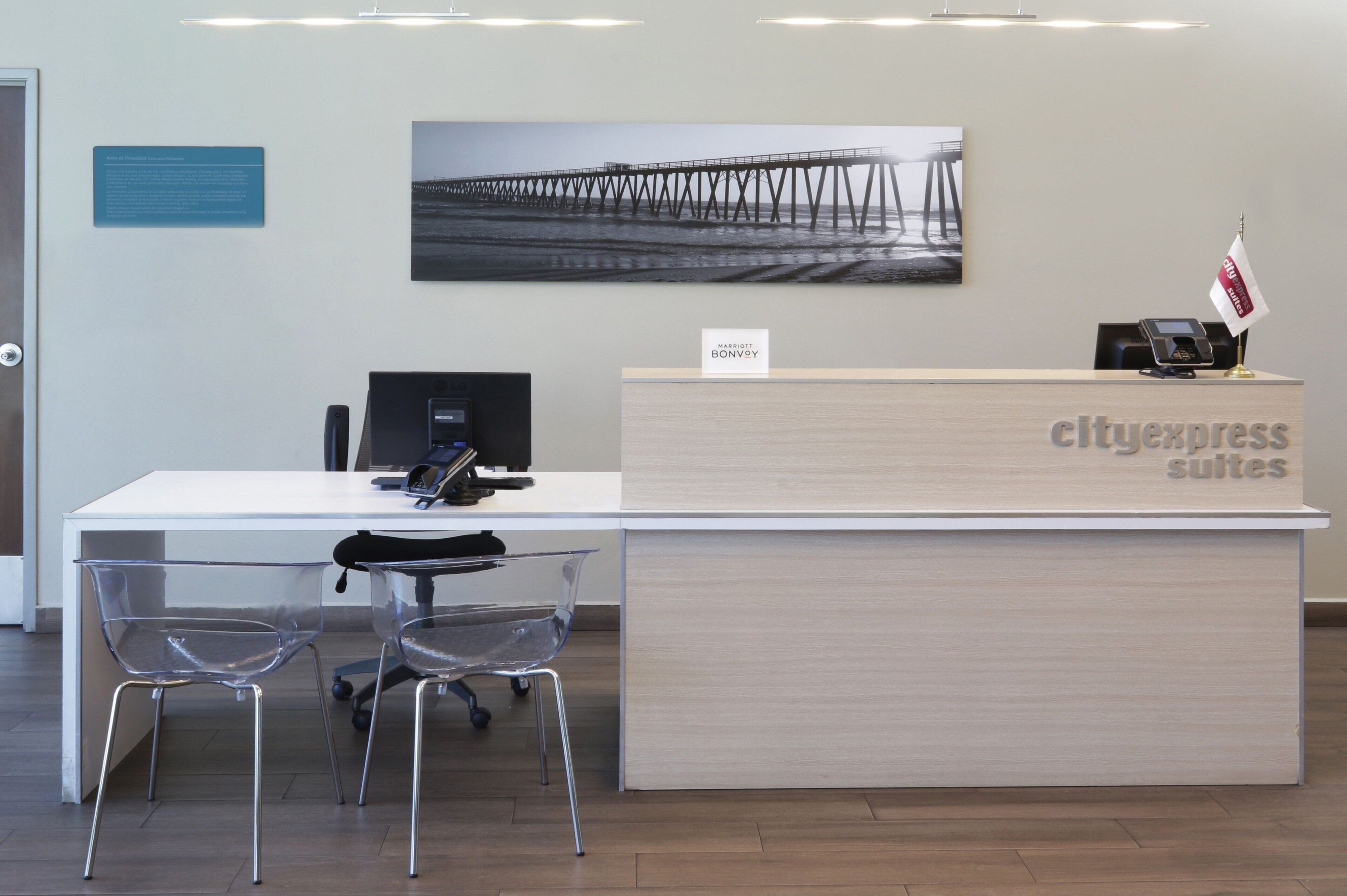 A hotel reception desk with a sign reading "cityexpress suites," a computer, and two transparent chairs.