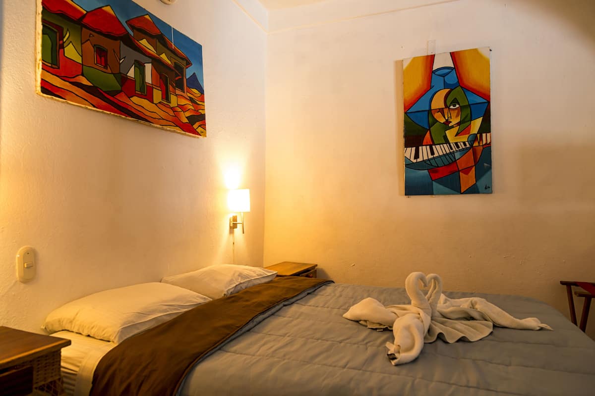 A hotel room with a bed, pillows, a blanket, a wall-mounted lamp, and two paintings.