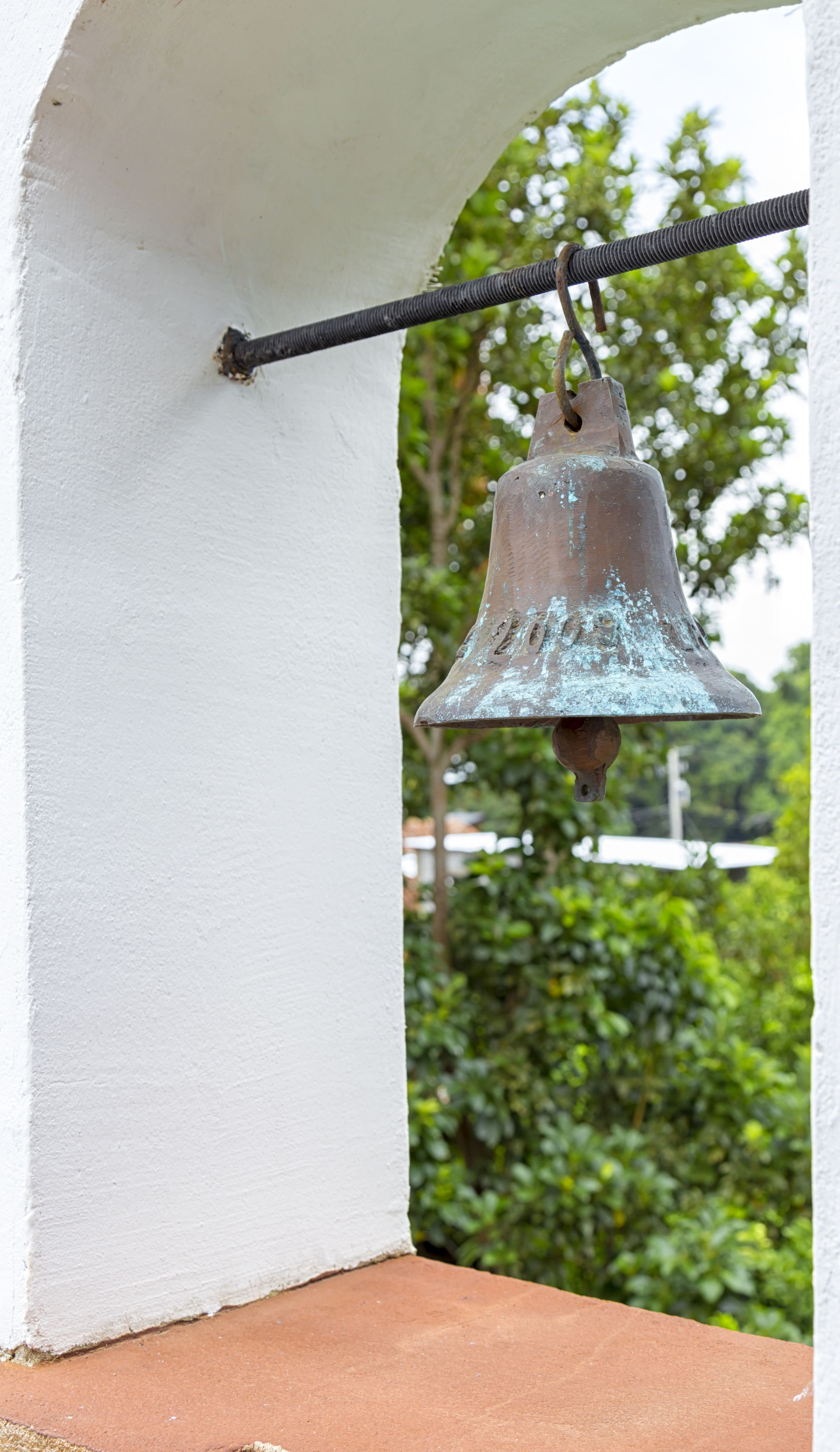 A weathered metal bell hanging from a black metal rod attached to a white wall.
