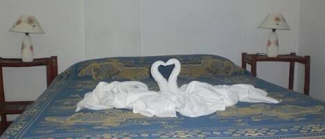 Una cama con colcha de diseño azul y dorado, dos mesitas de noche con lámparas y decoración de toallas con forma de cisne y ganso sobre la cama.