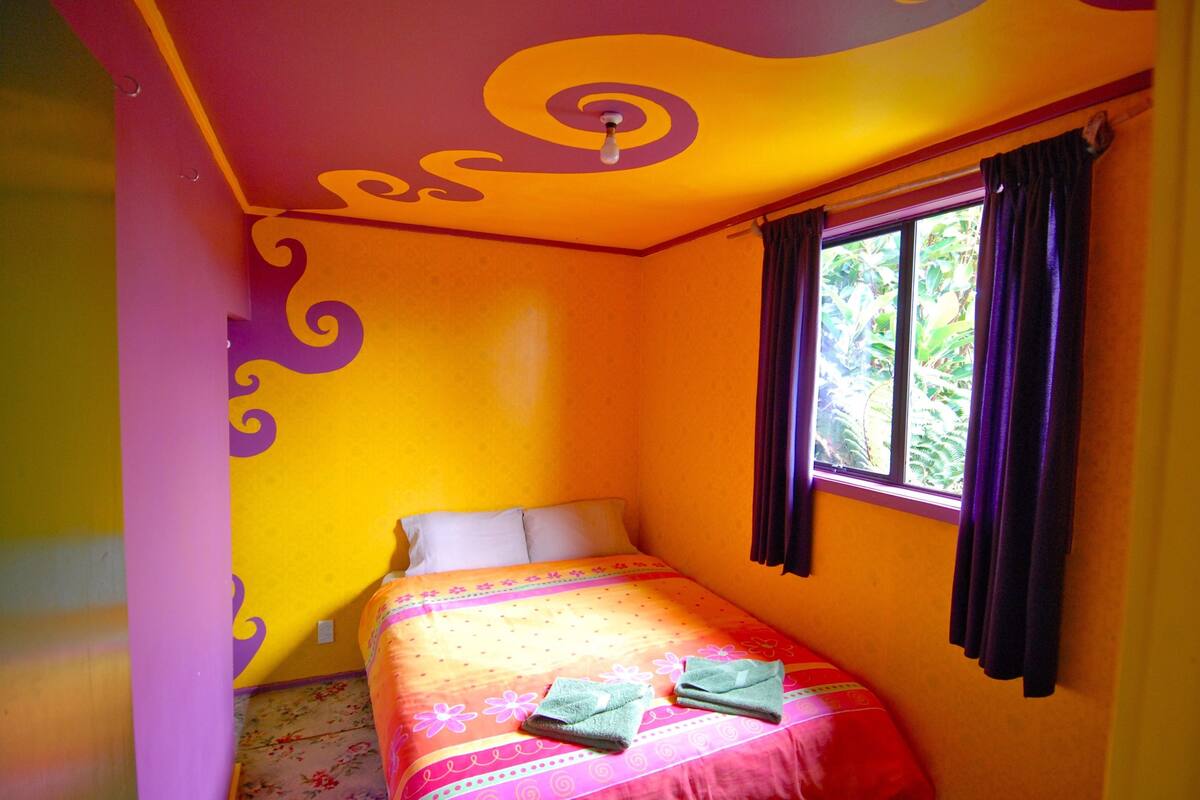 A small, brightly colored room with a single bed, a window with curtains, and a ceiling with a swirl design.