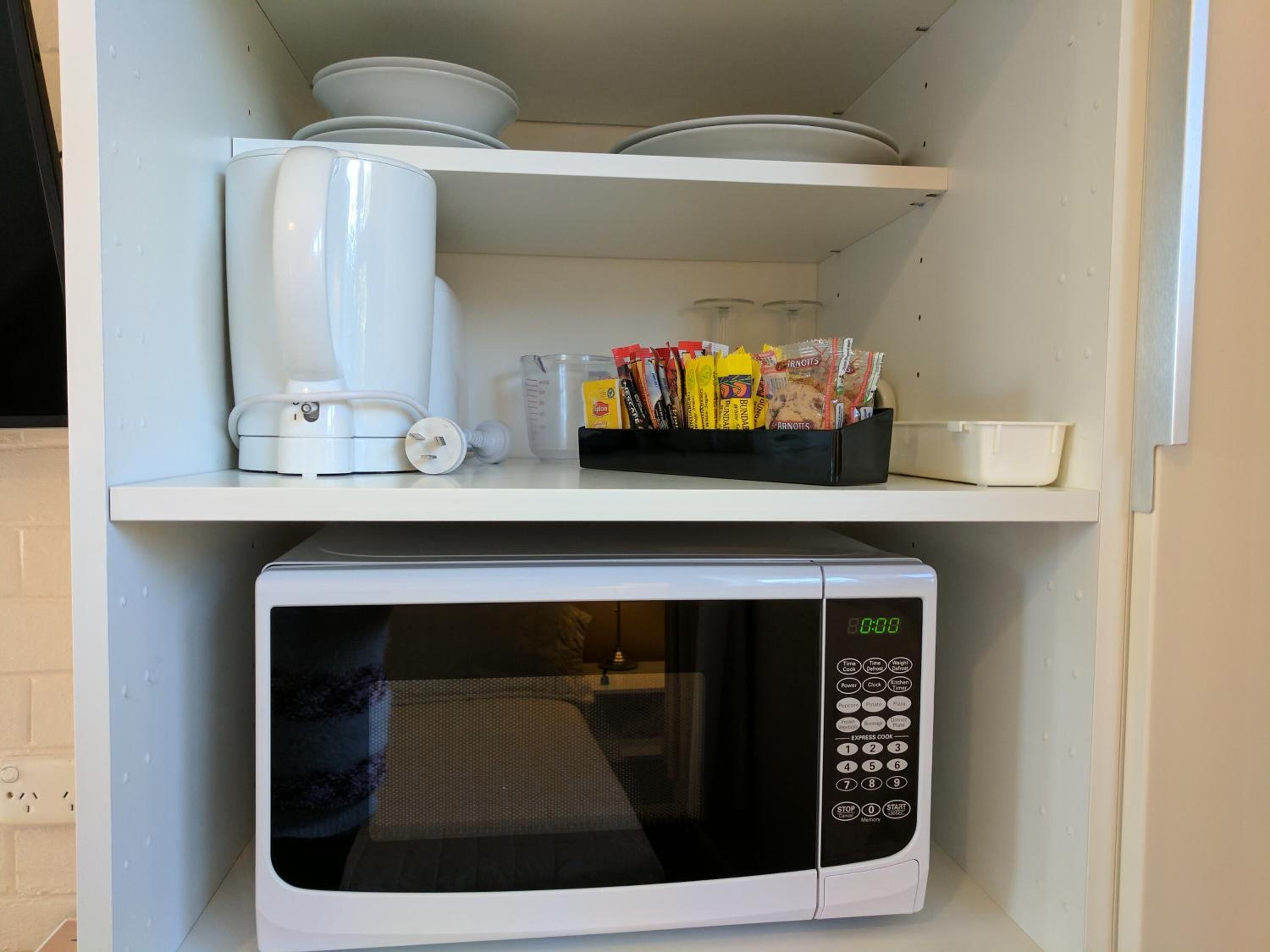 A kitchen with a microwave and a coffee maker on the countertop, and a shelf above containing bowls, a measuring cup, and snack packets.