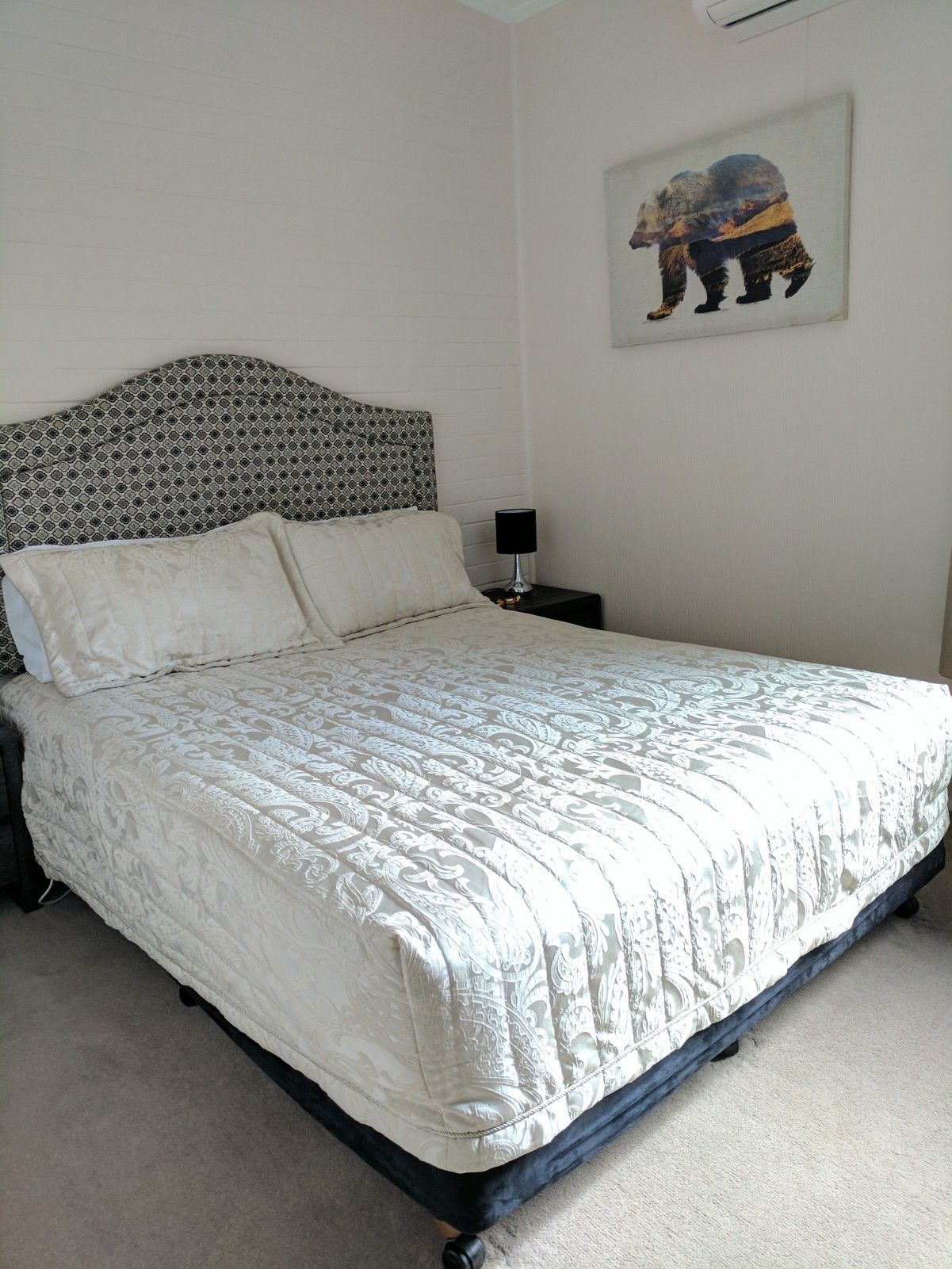 A neatly made bed with a patterned bedspread and pillows, a bedside table with a lamp, and a wall-mounted picture of a bear.