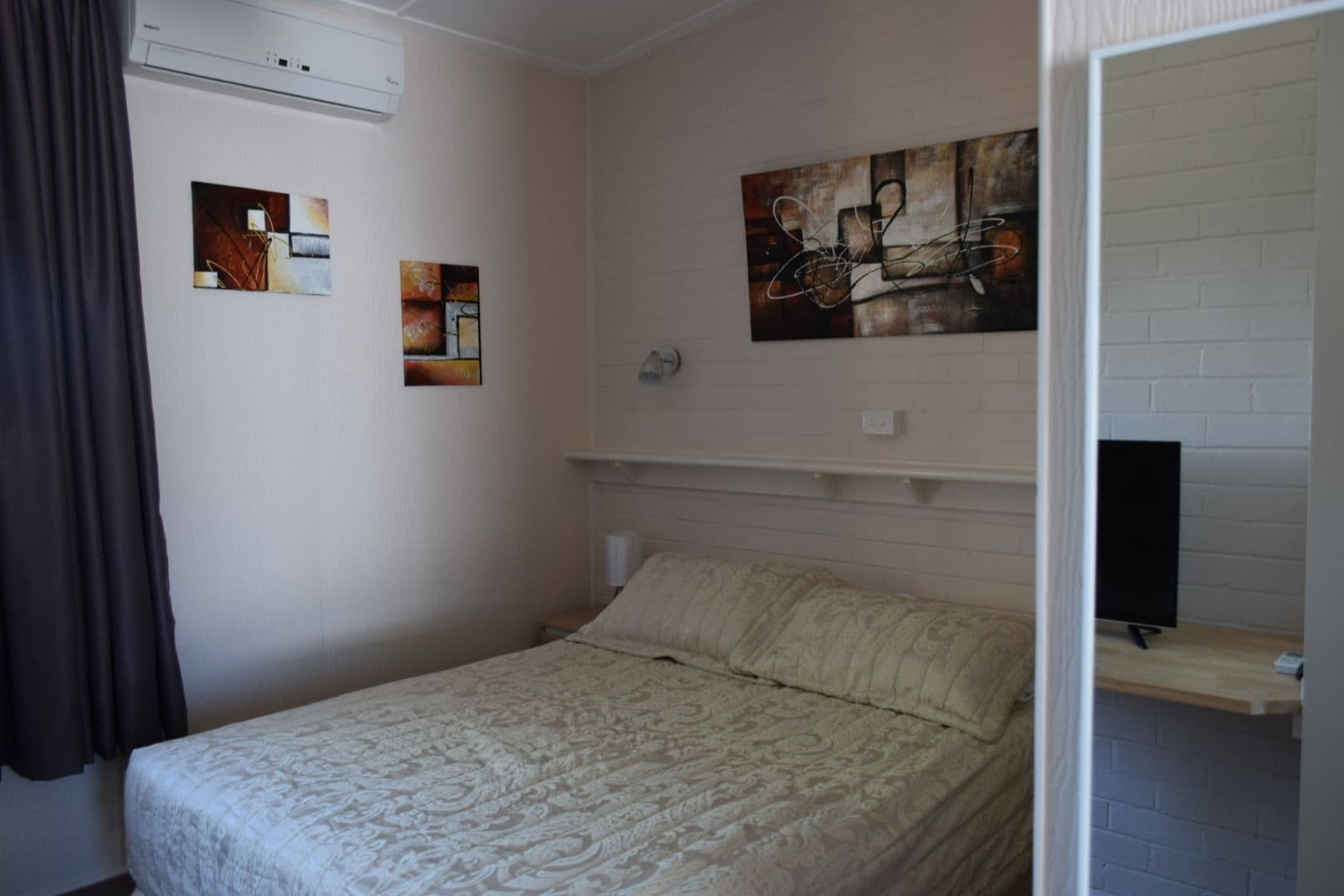 Un dormitorio con cama, cabecera blanca, estante con televisor y dos cuadros en la pared.