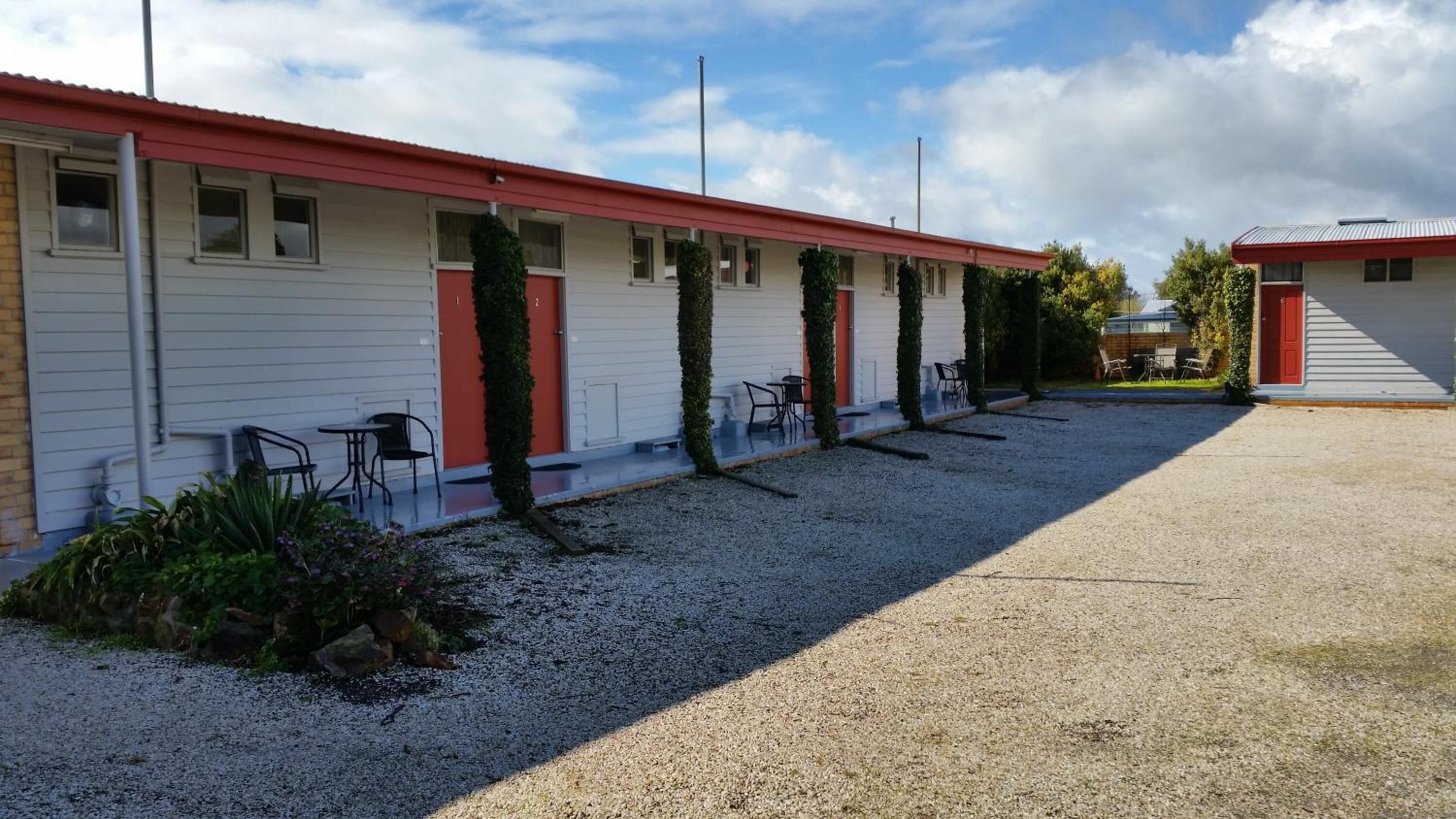 Un motel avec un toit rouge et un revêtement blanc, des places assises à l’extérieur et une allée en gravier.