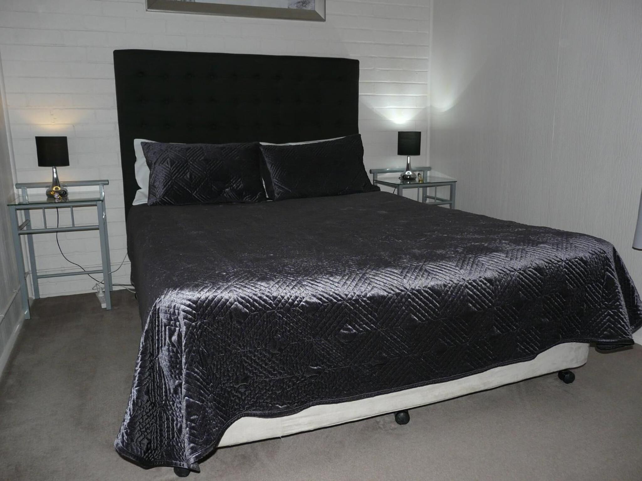 A neatly made bed with a black quilt, two bedside tables with lamps, and a white brick wall.