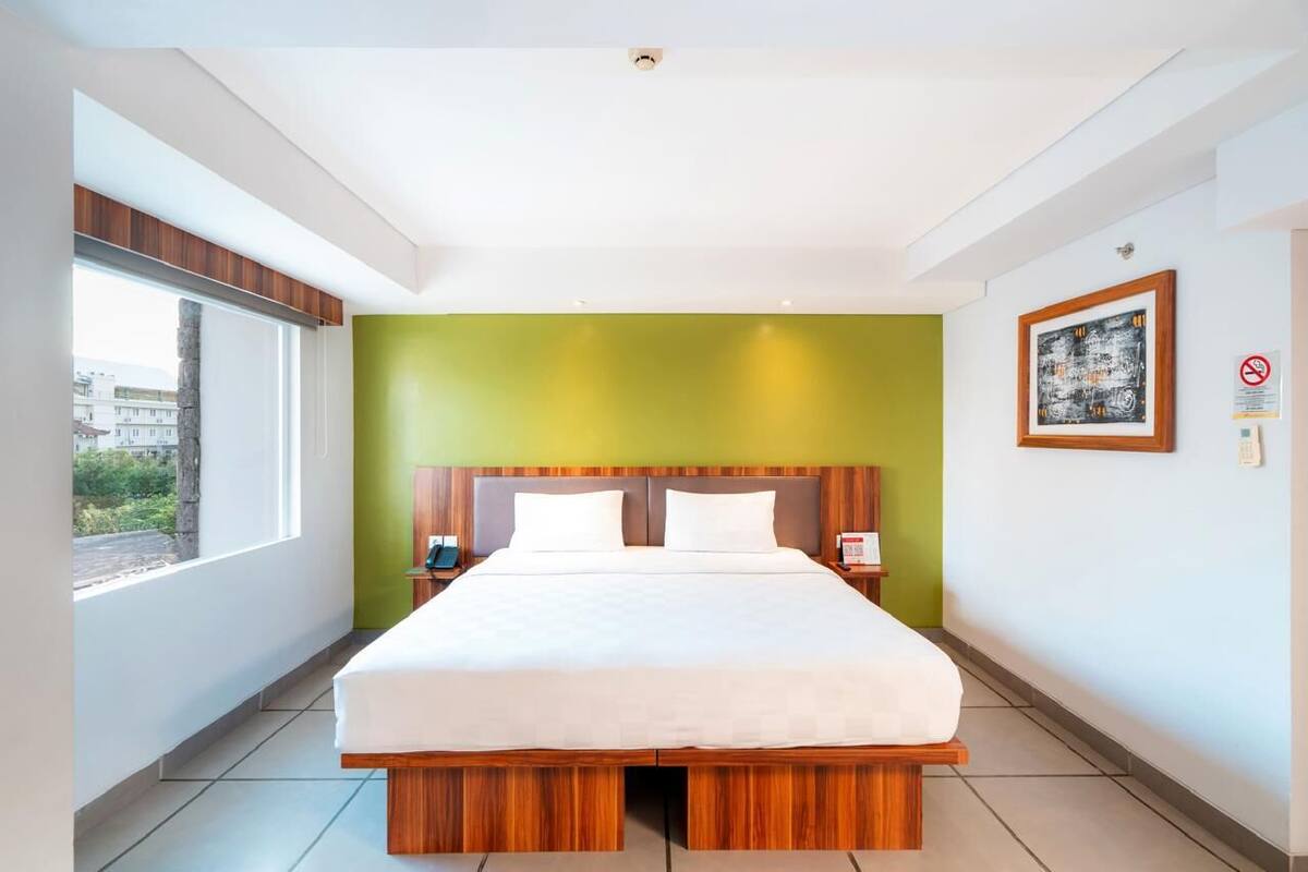 A modern hotel room with a large bed, a green accent wall, and a window providing a view of the outside.