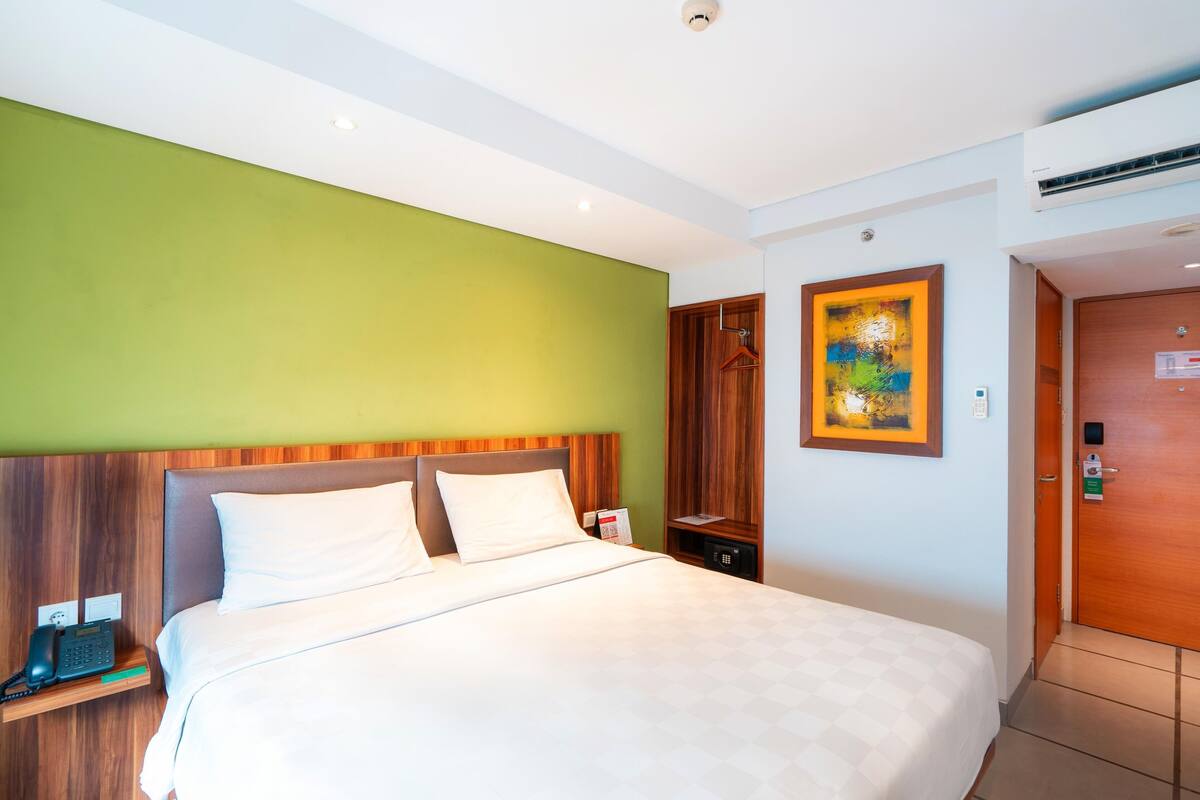 A hotel room with a large bed, a wooden headboard, a bedside phone, a small table, a wardrobe, and a painting on the wall.