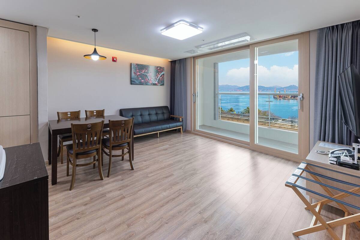 A modern hotel room with a dining area, a sofa, and a large window offering a view of the sea.