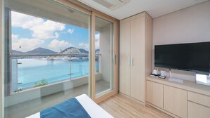 A modern hotel room with a balcony, a large TV, and a view of a coastal city. - The Hotel Soo (Yeosu)