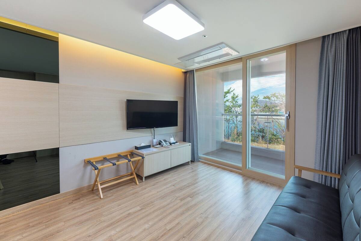 A modern living room with a flat-screen TV, a wooden desk, a cushioned sofa, and a balcony with a view of the outdoors.