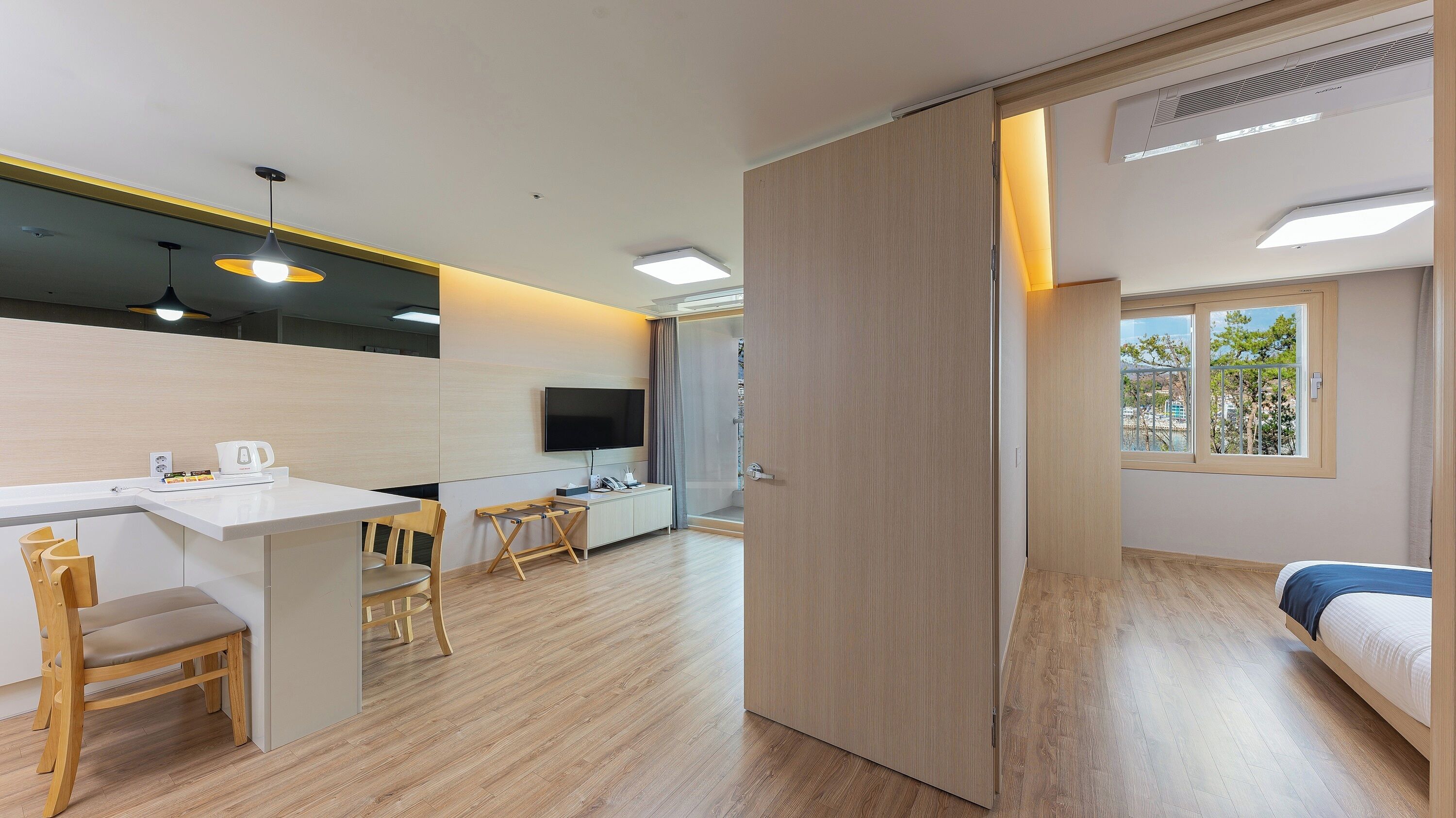 A modern hotel room with a wooden floor, a desk with a chair, a bed, a TV mounted on the wall, and a sliding door leading to a shower area.