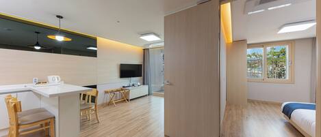 A modern hotel room with a wooden floor, a desk with a chair, a bed, a TV mounted on the wall, and a sliding door leading to a shower area.