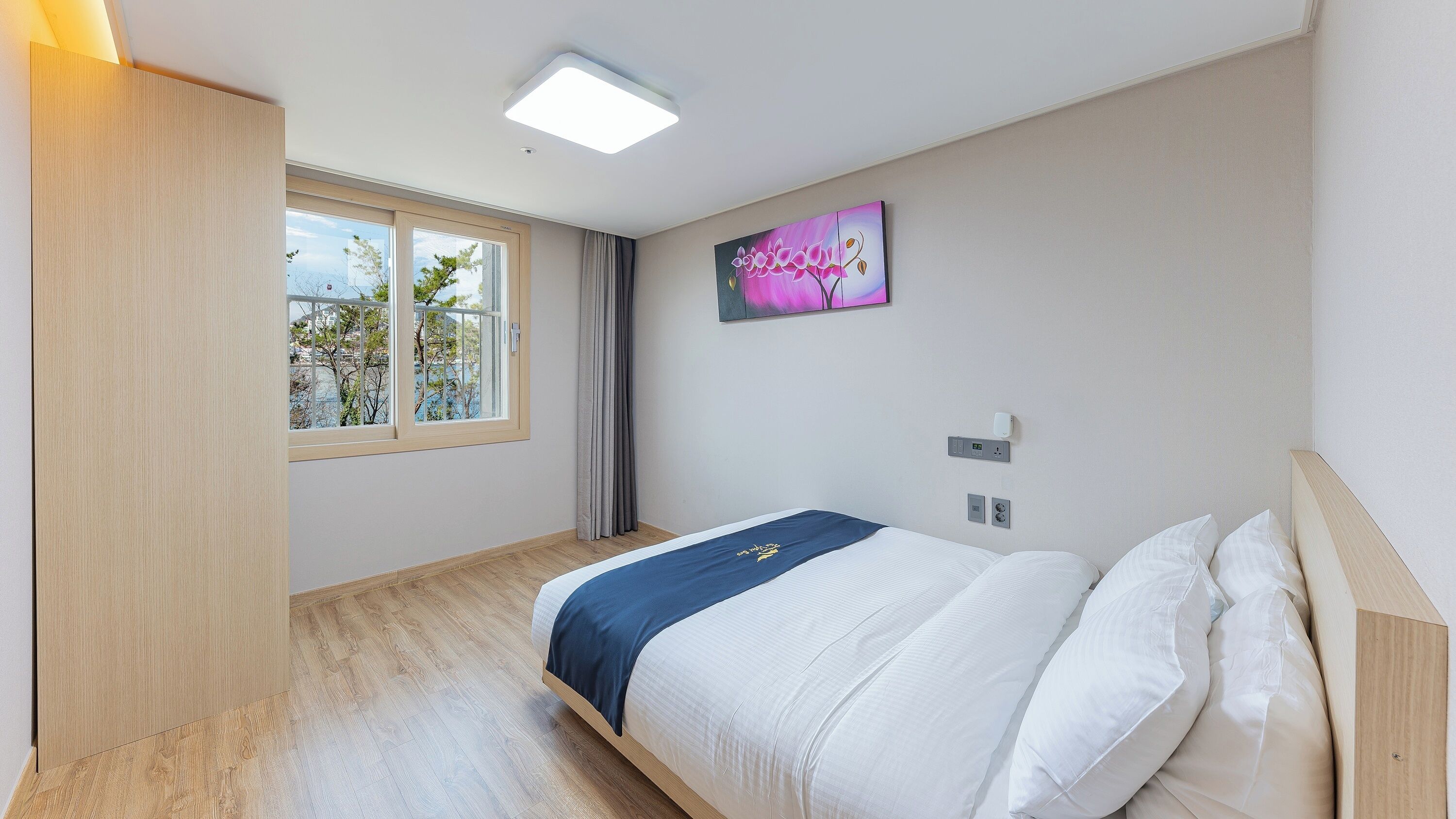 Un dormitorio con cama, ventana con vistas, un cuadro en la pared y una lámpara en el techo.