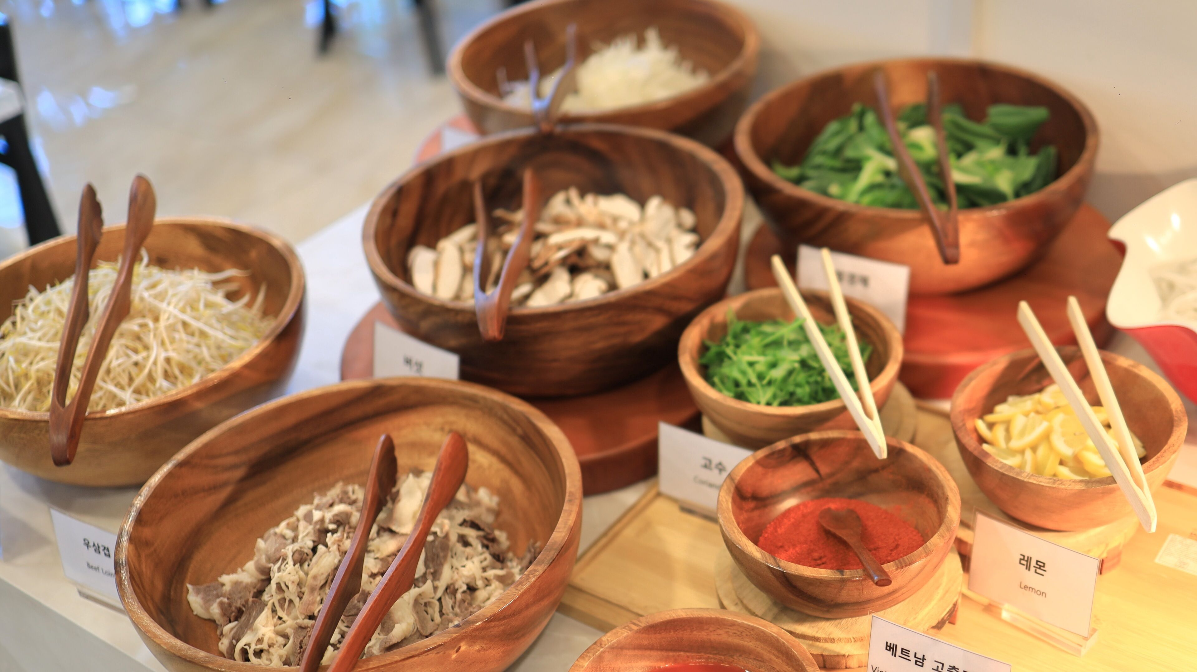 Diversos cuencos de madera con diferentes alimentos, cada uno etiquetado con un nombre en un alfabeto extranjero.
