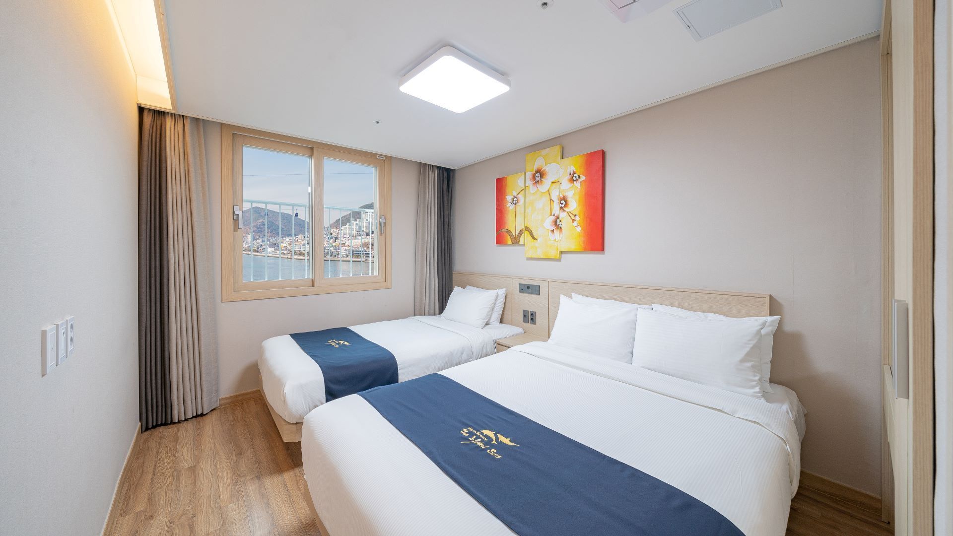 A hotel room with two beds, a city view, a painting on the wall, and a ceiling light.