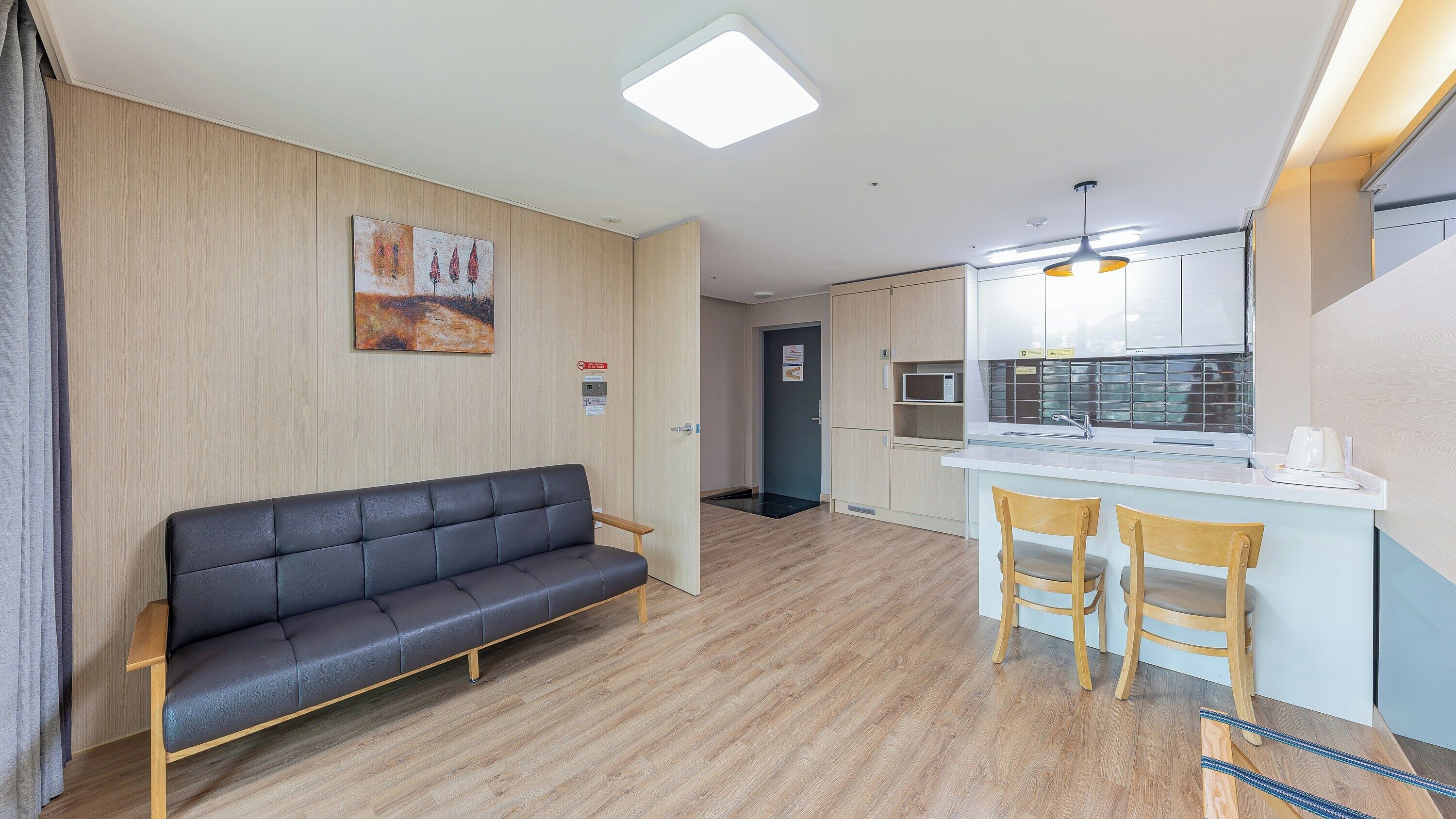 A modern hotel room with a sofa, a small kitchen area with a microwave, and a wooden floor.