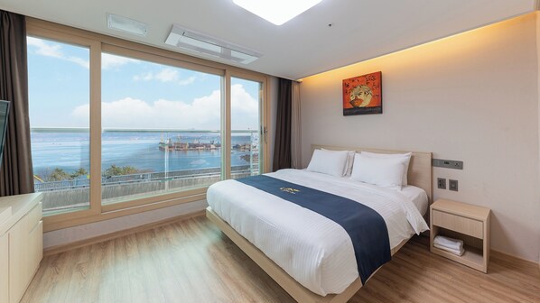 A hotel room with a large window offering a view of the sea and port, a bed with white linens, a nightstand, and a painting on the wall. - The Hotel Soo (Yeosu)