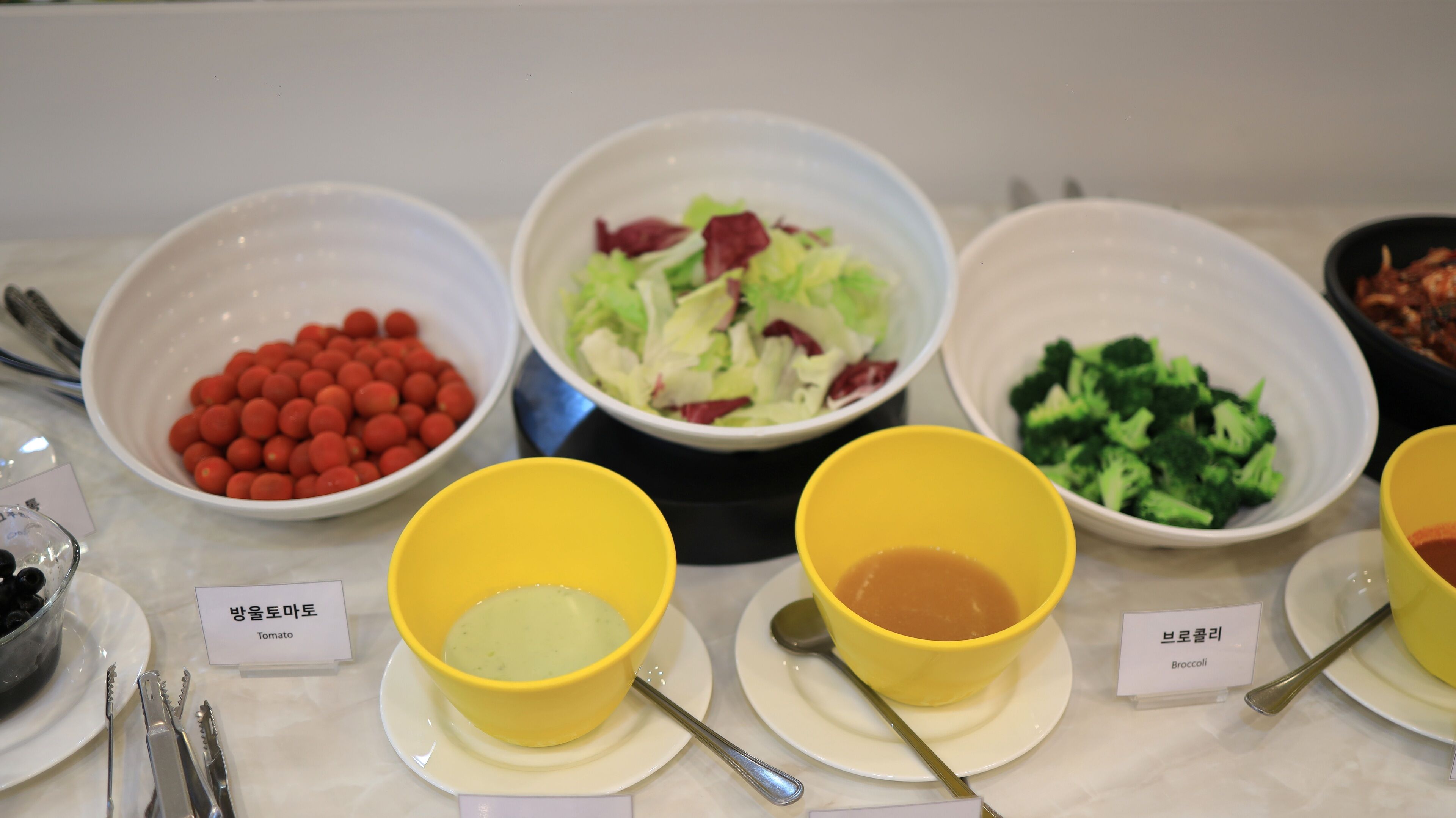 Una mesa con diferentes alimentos y recipientes etiquetados, incluyendo tomates, ensalada y brócoli.