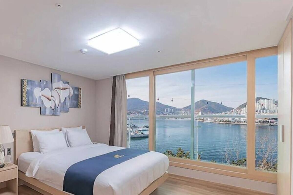 A hotel room with a bed, a nightstand, a lamp, a large window with a city and water view, and a painting on the wall.