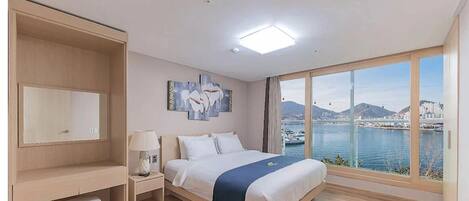 Un cuarto de hotel con una cama, una mesita de noche, una lámpara, un ventanal grande con vista a la ciudad y al agua, y un cuadro en la pared.