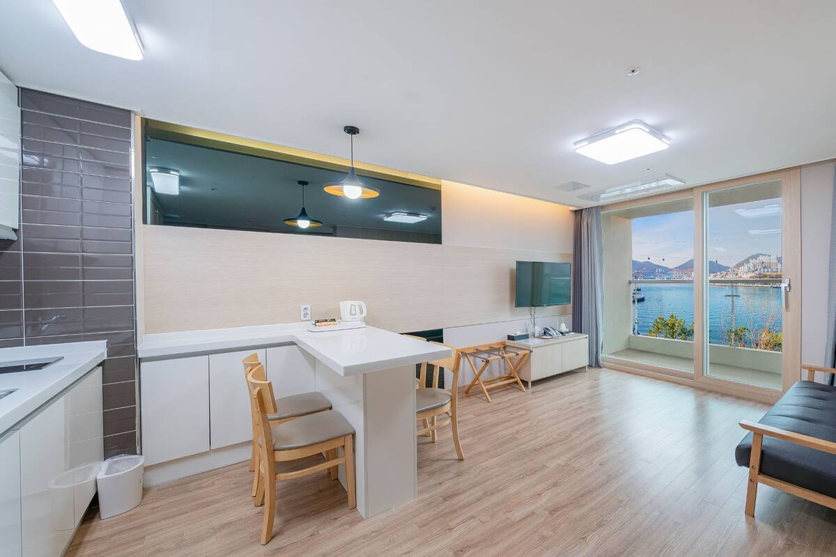 A modern kitchen with a dining area, a sofa, and a large window with a view of a body of water and mountains.