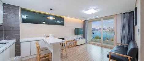 A modern kitchen with a dining area, a sofa, and a large window with a view of a body of water and mountains.