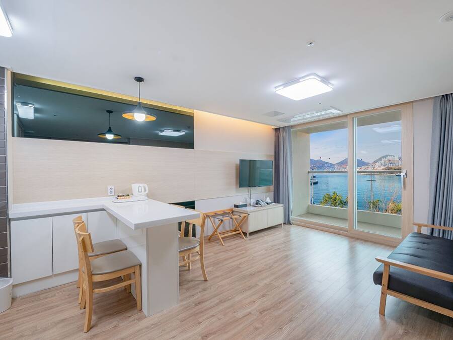 A modern kitchen with a dining area, a sofa, and a large window with a view of a body of water and mountains.