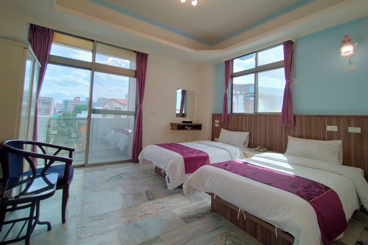 A hotel room with two beds, a desk, and a chair. There are large windows with curtains, a ceiling light fixture, and a view of the city.