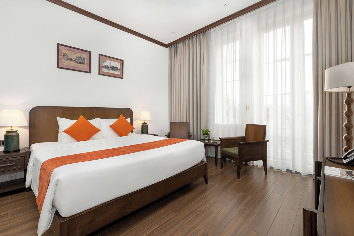 Habitación de hotel con una cama grande, dos mesitas de noche con lámparas, un escritorio con silla y un ventanal con cortinas.