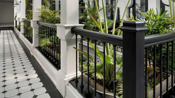 Un pasillo con suelo de baldosas geométricas, columnas blancas y grandes plantas verdes.