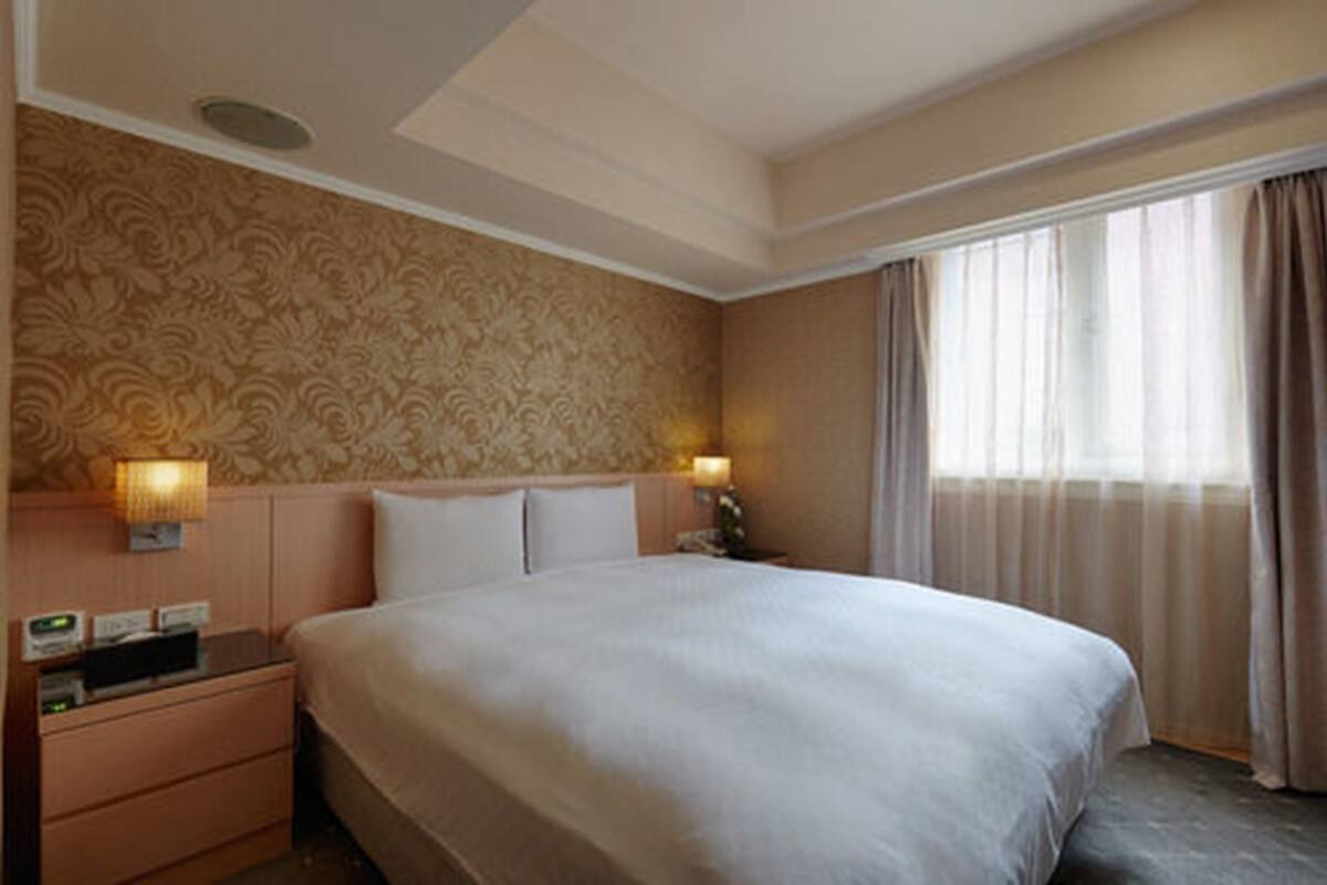 A hotel room with a large bed, two bedside tables with lamps, a patterned wallpaper, and a window with curtains.