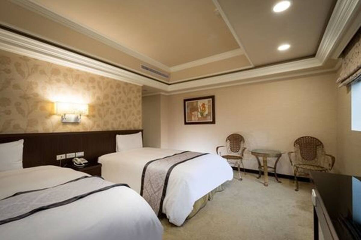 A hotel room with two beds, a small table, two chairs, a TV, and a painting on the wall.