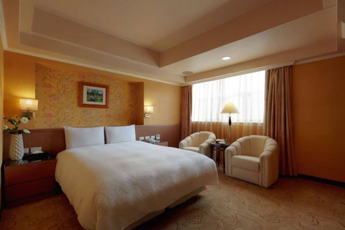 A hotel room with a large bed, two armchairs, a small table, a nightstand, a lamp, a picture on the wall, and a window with curtains.
