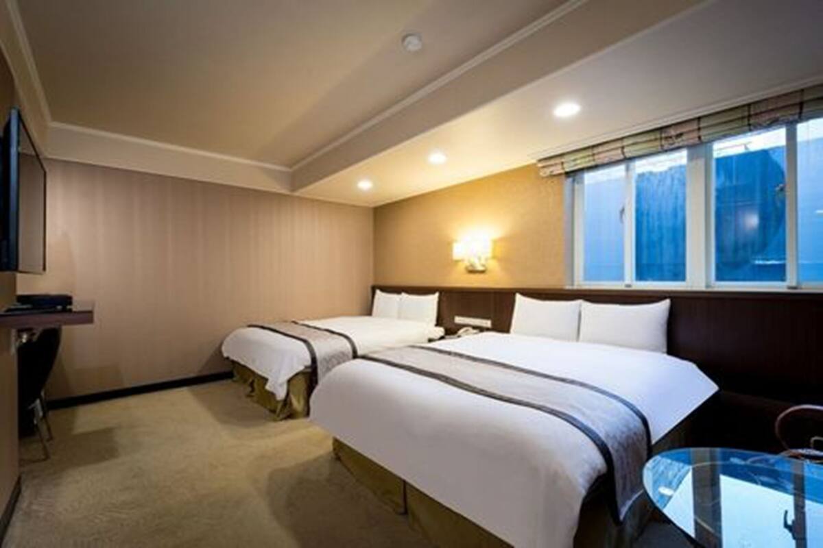 A hotel room with two beds, a bedside table, a chair, a small table, and a window with blinds.